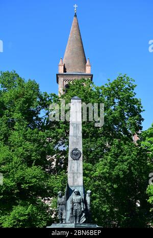 12 mai 2020, Brandebourg, Potsdam: Le mémorial sur le cimetière soviétique d'honneur sur la Bassinplatz, en arrière-plan derrière les arbres, le sommet de l'église paroissiale catholique Saint-Pierre et Paul peut être vu. Environ 400 tombes et un obélisque décoré de figures de bronze commémorent les soldats tombés de l'armée soviétique pendant la bataille de Berlin. Les soldats de l'Armée rouge qui se tiennent sur le piédestal du monument en grès et en granit représentent les quatre branches de l'armée par un soldat de garde, un pilote de char, un fantassin naval et un pilote. Le cimetière d'honneur est sous protection monumentale depuis 1987. Banque D'Images