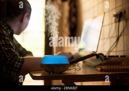 La vapeur qui s'échappe de l'huile essentielle diffuse avec une LED bleue pendant que la femme travaille sur un ordinateur portable. Banque D'Images