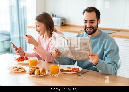 Portrait de jeune couple joyeux utilisant le téléphone portable et lisant le journal tout en prenant le petit déjeuner dans la cuisine confortable à la maison Banque D'Images