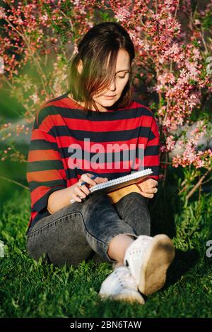 Une femme assise sur l'herbe dans un jardin fleuri travaille avec un bloc-notes et un crayon Banque D'Images