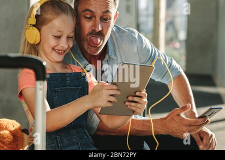 Jolie fille portant un casque regardant une tablette numérique avec son père collant hors de la langue. Père et fille faisant des vidéos amusantes sur tablette numérique Banque D'Images