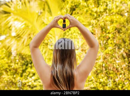 Femme tient dans les mains la bouteille d'huile essentielle sur fond jaune naturel, vue de l'arrière. Banque D'Images