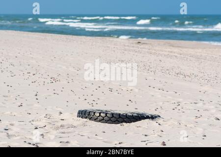 Grand pneu en caoutchouc noir laissé sur une plage de sable avec mer bleue et vagues sur fond, environnement pollution concept Banque D'Images