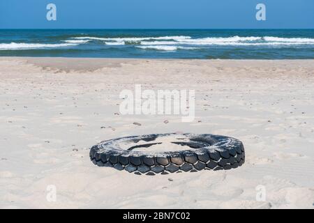 Grand pneu en caoutchouc noir laissé sur une plage de sable avec mer bleue et vagues sur fond, environnement pollution concept Banque D'Images