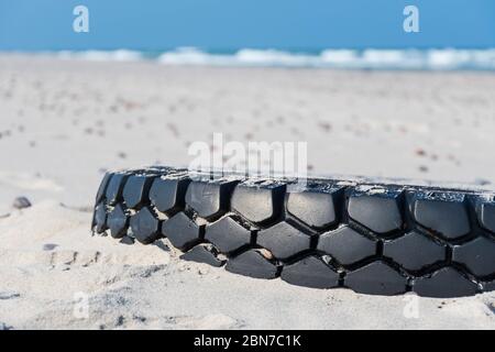 Grand pneu en caoutchouc noir laissé sur une plage de sable avec mer bleue et vagues sur fond, environnement pollution concept Banque D'Images
