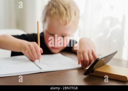 Petit garçon triste et fatigué apprenant à la maison. Regarder un webinaire sur mobile, écrire sur un ordinateur portable Banque D'Images