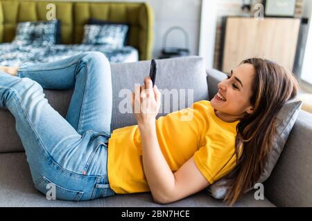 Jolie femme sourire comme elle lit un message sms sur son mobile tout en vous relaxant pieds nus sur un canapé dans son salon Banque D'Images