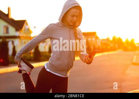 Jeune femme coureur, athlète s'étirant, jogging dans la rue de la ville au soleil de printemps. Belle femme caucasienne de formation, à l'écoute de la musique. Concept de sport, mode de vie sain, mouvement, activité. Banque D'Images