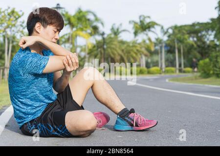 Homme asiatique se servir des mains pour tenir son coude tout en courant sur la route dans le parc. Blessure par rapport à l'entraînement. Banque D'Images