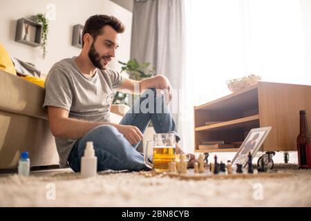 Jeune homme positif barbu assis sur le sol avec une tasse à bière et jouant aux échecs avec un ami à distance pendant l'isolement à la maison Banque D'Images