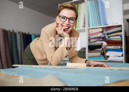 Portrait de jeune tailleur de lunettes souriant à l'appareil photo tout en faisant le motif sur le tissu à la table Banque D'Images