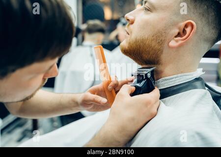 Barbier tailler homme barbu avec machine à raser dans le barbershop. Processus de coiffure. Gros plan d'un stylist coupant la barbe d'un homme barbu. Banque D'Images