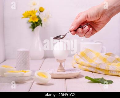 La main femelle fait un œuf bouilli avec une cuillère sur un support sur fond de bois blanc. Le concept de petit déjeuner, matin d'été. Régime alimentaire sain facile. Pour Banque D'Images