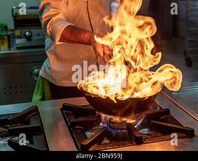 Chef cuisinez avec feu dans une poêle. Chef professionnel dans une cuisine commerciale de style flambe. Cuisiniers friture des aliments dans une poêle à frire sur une table de cuisson à gaz en co Banque D'Images