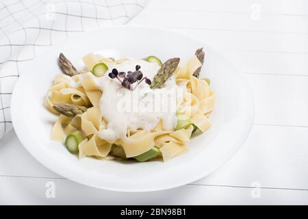 Plat de pâtes maison frais de fettuccine ou tagliatelle, asperges vertes, sauce blanche, dans une assiette blanche sur table en bois blanche Banque D'Images