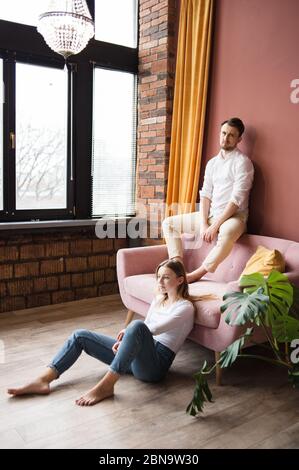 L'homme est assis sur le dos du canapé et regarde la caméra, la femme est assise sur le sol près. Banque D'Images