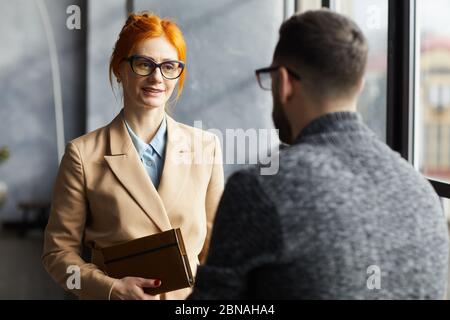 Femme d'affaires aux cheveux rouges en lunettes de vue parlant à l'homme d'affaires pendant qu'ils se tiennent dans le couloir du bureau Banque D'Images