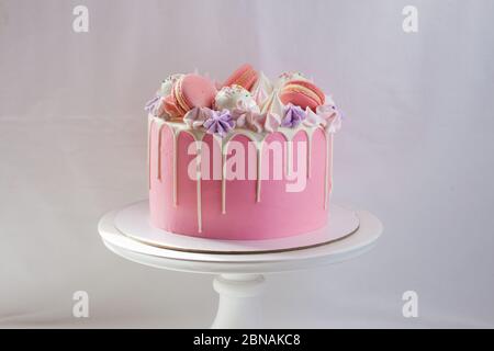 Gâteau rose tendre décoré de chocolat blanc fondu, macarons, meringues, pâtes à gâteau et bonbons sur cakestan blanc. Arrière-plan simple. Banque D'Images