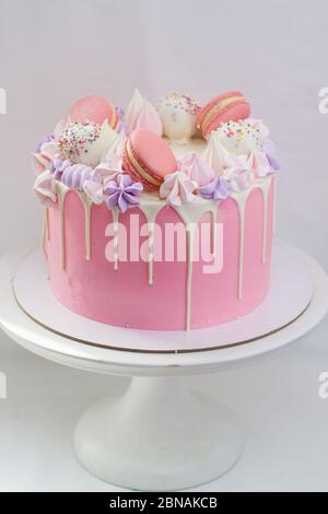 Gâteau rose tendre décoré de chocolat blanc fondu, macarons, meringues, pâtes à gâteau et bonbons sur cakestan blanc. Arrière-plan simple. Banque D'Images