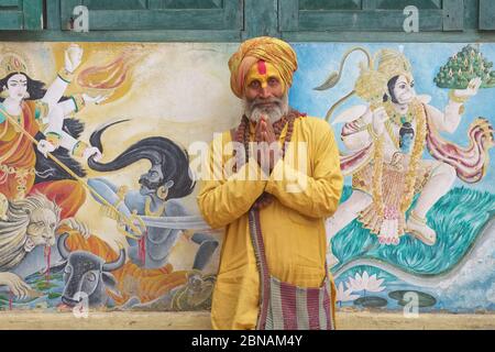 Un hindou sadhu (Saint-homme) qui exécute Namaste (salutation), Temple de Pashupatinath, Katmandou, Népal, entre les peintures murales de la déesse Durga et dieu singe Hanuman Banque D'Images