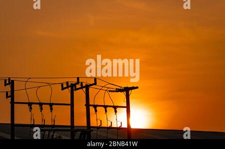 Alimentation électrique triphasée pour le transfert de puissance par réseaux électriques. Poteaux électriques haute tension dans l'usine et le toit du bâtiment et ciel orangé de coucher de soleil. Banque D'Images