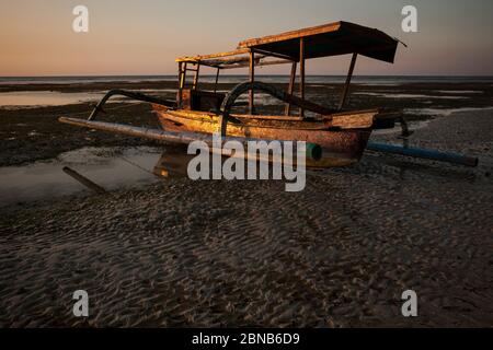 Vue horizontale au coucher du soleil d'un vieux et rouillé traditionnel indonésien outrigger bateau de pêche s'est échoué sur la plage en raison de marée basse, Gili Air, îles Gili Banque D'Images