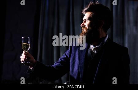 beau barbu avec moustache à cheveux et barbe longue et visage sérieux debout dans un costume bleu tenant un verre de whisky à main levée Banque D'Images