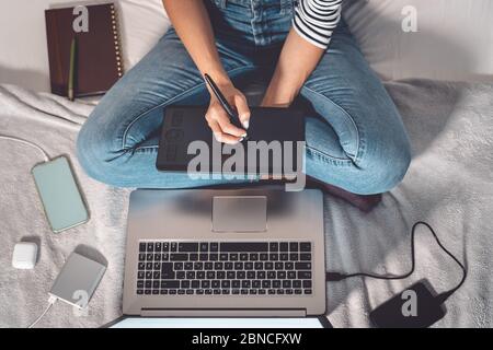 Graphiste femme intelligente travaillant sur ordinateur à la maison - jeune femme dessin avec stylo interactif et ordinateur portable dans le lit Banque D'Images