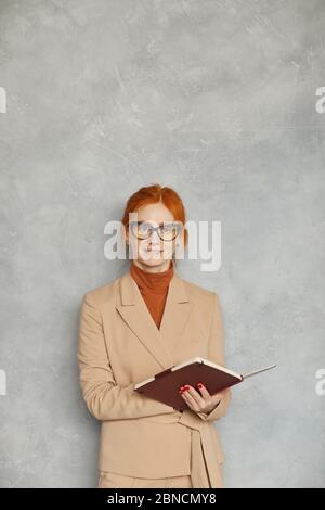Portrait d'une femme d'affaires mûre aux cheveux rouges dans des lunettes tenant son bloc-notes et souriant à l'appareil contre le mur gris Banque D'Images