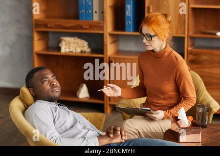Psychologue mature assis sur un fauteuil et parlant à un homme africain alors qu'il est allongé sur le canapé au bureau Banque D'Images