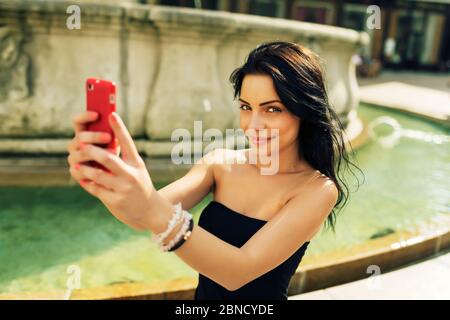 Belle fille souriant et prenant selfie. Jeune jolie femme s'amusant et prenant des photos avec un smartphone rouge devant une fontaine dans l'histoire Banque D'Images