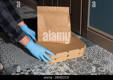 Vue rognée de l'homme de livraison dans des gants en latex mettant des boîtes à pizza et des paquets sur le sol près de la porte Banque D'Images