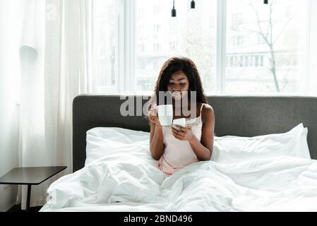 Femme afro-américaine tenant une tasse de thé et un smartphone sur le lit dans la chambre Banque D'Images