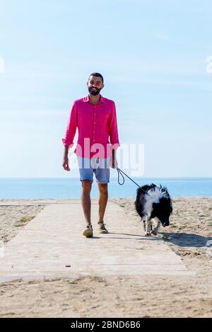 Jeune homme marchant avec son chien attaché sur la plage Banque D'Images