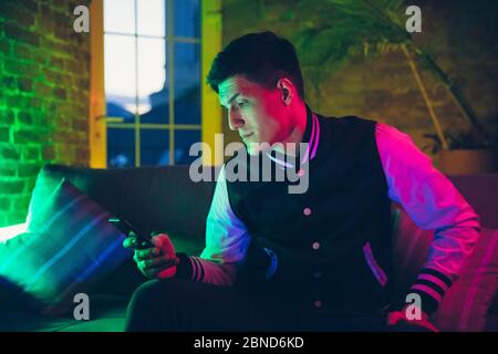 Calme. Portrait cinématographique d'un homme élégant dans un intérieur éclairé au néon. Tons comme les effets de cinéma, couleurs vives. Modèle caucasien utilisant des gadgets, des dispositifs dans des lumières colorées à l'intérieur. Culture de la jeunesse. Banque D'Images