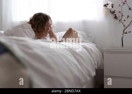 Femme utilisant une tablette informatique couchée sur un lit dans une chambre blanche. Banque D'Images