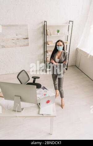 vue en grand angle de la femme d'affaires en tenue formelle et masque médical debout avec bras croisés près de la table avec des ordinateurs à la maison Banque D'Images