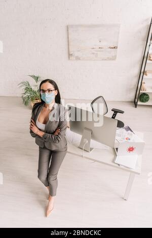 vue en grand angle de la femme d'affaires en tenue formelle et masque médical debout avec bras croisés près de la table avec des ordinateurs à la maison Banque D'Images