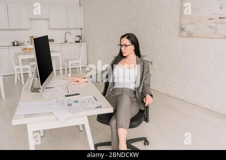 femme d'affaires attirante tenue habillée à la table avec moniteur d'ordinateur et documents dans la cuisine Banque D'Images