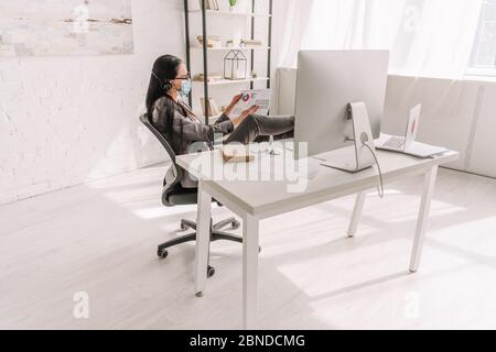 femme d'affaires en tenue officielle et masque médical regardant les papiers près de la table avec moniteur d'ordinateur pendant le travail à la maison Banque D'Images