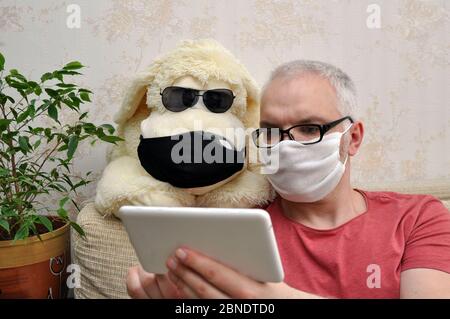 Un homme dans un masque médical à côté d'un chien jouet, qui est à la maison sur l'auto-isolement, regarde un film. Concept de prévention des coronavirus Banque D'Images