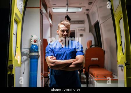 Un bel homme dans un uniforme médical regardant la caméra avec un sourire, les bras repliés, voiture d'ambulance en arrière-plan Banque D'Images