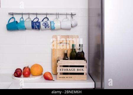 Les tasses en céramique multicolore aux tons bleus sont accrochées aux crochets au-dessus de la table de cuisine. Les bouteilles d'huile d'olive sont dans une boîte en bois. Poires, pommes et vigne Banque D'Images