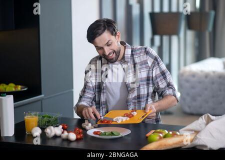 Jeune homme barbu versant des champignons et des tomates hachés. Banque D'Images