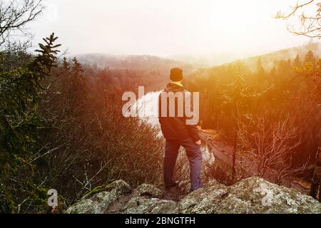 Jeune homme en veste, pantalon et sac à dos debout sur le rocher regardant la rivière Vltava et la vallée au coucher du soleil. Banque D'Images