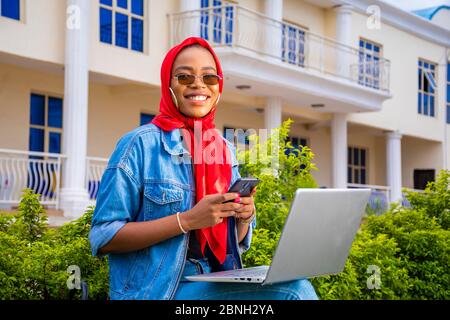 belle jeune femme noire assise seule avec son ordinateur portable et tenant son téléphone à l'extérieur dans un parc souriant et se sentant excitée Banque D'Images