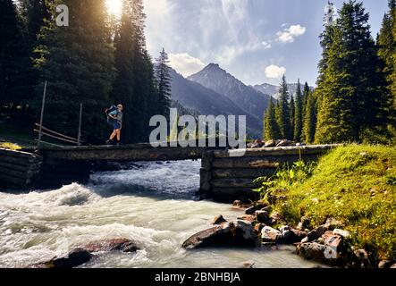 Touriste avec grand sac à dos vert traverse la rivière par le pont dans la vallée de la montagne dans le parc national de Karakol, Kirghizistan Banque D'Images