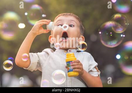 Un enfant à la bouche ouverte et aux yeux larges souffle des bulles de savon colorées dans la nature. Banque D'Images