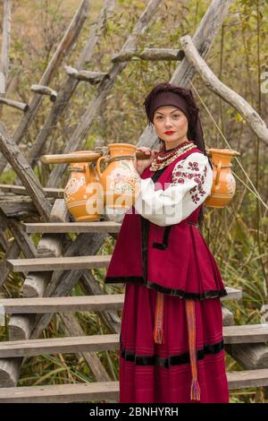 Une femme en vêtements traditionnels transporte de l'eau dans de vieux barils. Les vieux carafes sont portés par une fille dans un costume stylisé. Des vêtements originaux et magnifiques Banque D'Images