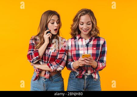 Image de deux jeunes filles belles portant des chemises à carreaux souriant et utilisant des téléphones cellulaires isolés sur fond jaune Banque D'Images